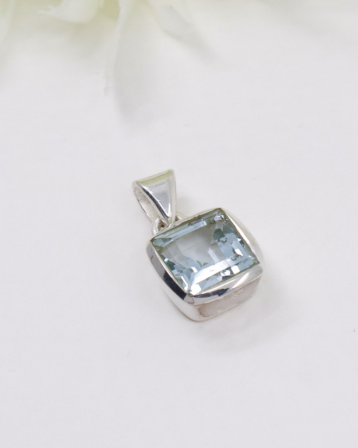 Faceted Blue Topaz sterling silver pendant with sparkling pale blue gemstone in a polished square setting photographed on a white background.