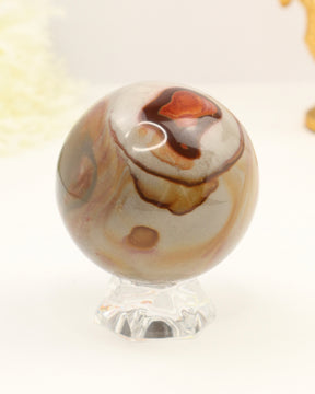 Polished Polychrome Jasper crystal sphere with natural earthy brown, cream and terracotta patterns displayed on a clear stand against a soft cream background.
