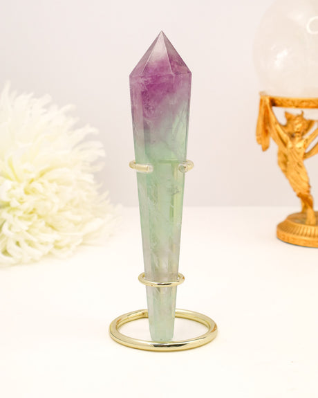 Polished Rainbow Fluorite crystal wand with purple and green banding displayed on a cream background with gold stand styling.